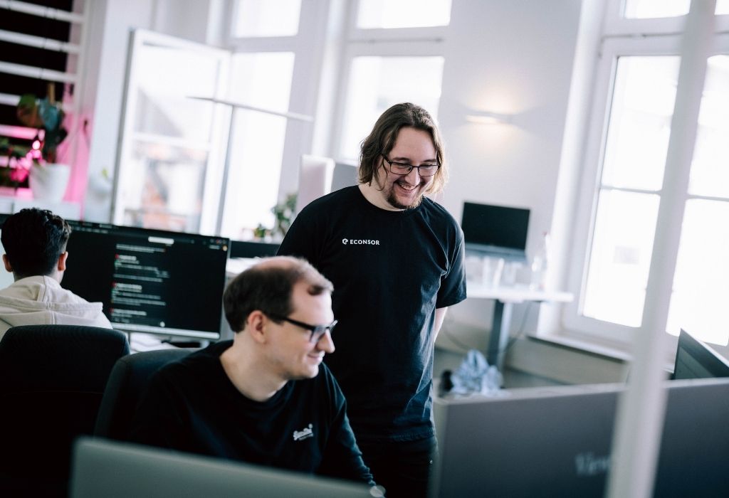 Zwei Entwickler lächeln und diskutieren gemeinsam an ihren Schreibtischen, während sie an einem Webentwicklungsprojekt arbeiten.